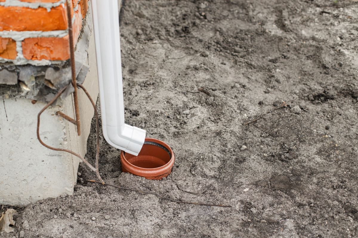White downspout emptying onto bare soil beside a brick foundation