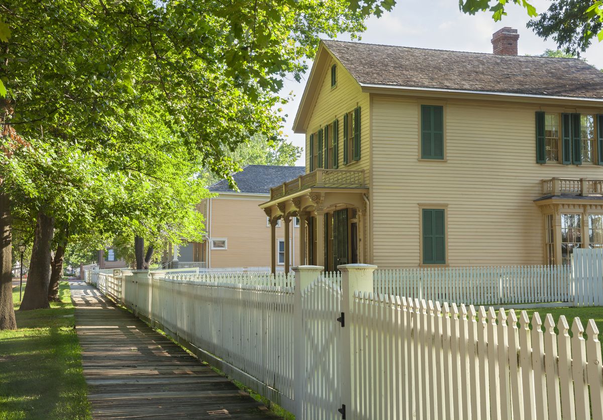 White picket fence with a gate beside a wooden sidewalk in front of a two-storey clapboard house